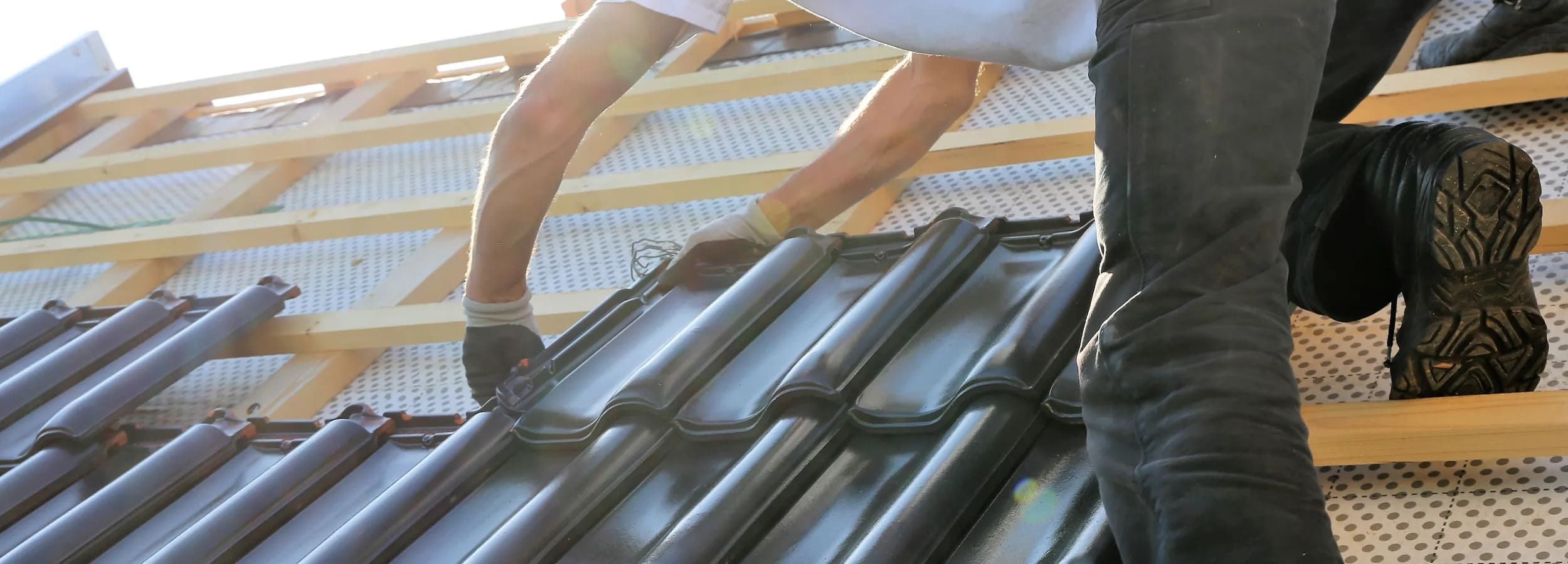 Person kniet auf einem Dach und verlegt schwarze Dachziegel, sichtbar sind Arme, Hände mit Handschuhen und Beine